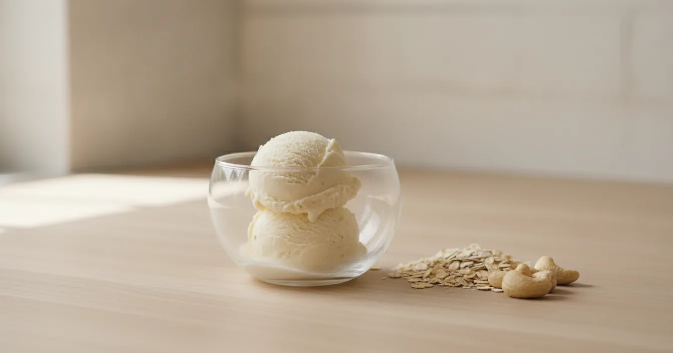 Scoops of pale dairy-free ice cream in a glass bowl on a wooden counter with rolled oats and cashews beside it.