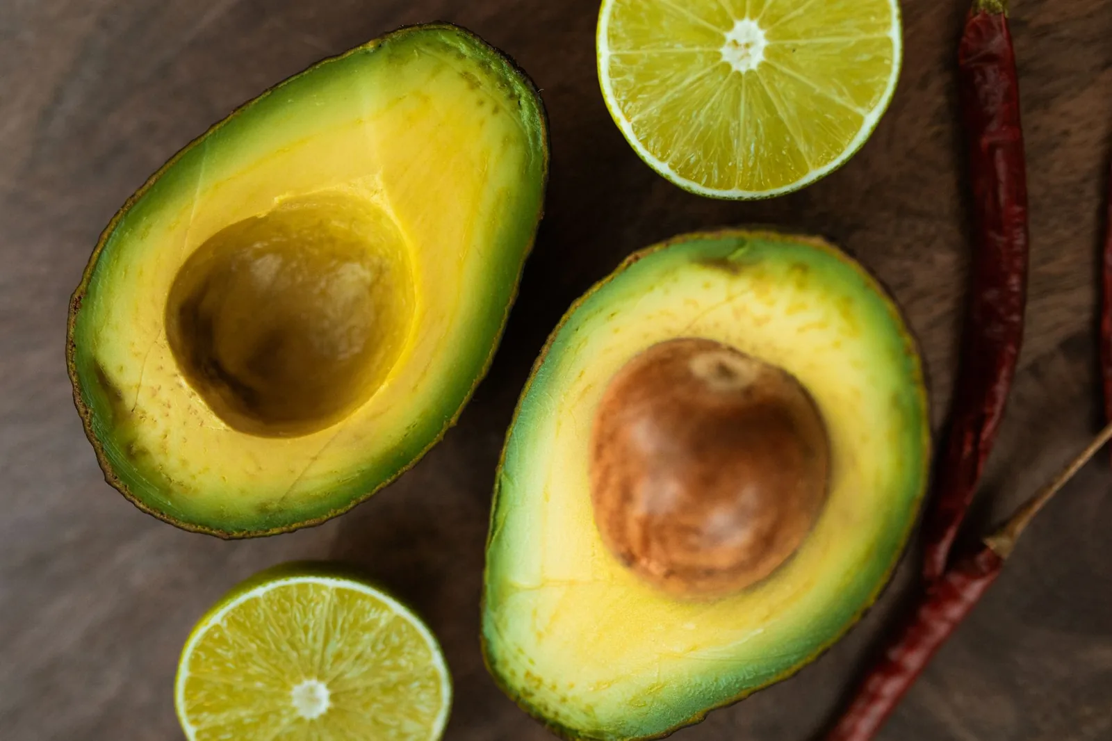 Two halved avocados with fresh limes and dried red chilies on a dark wooden board.