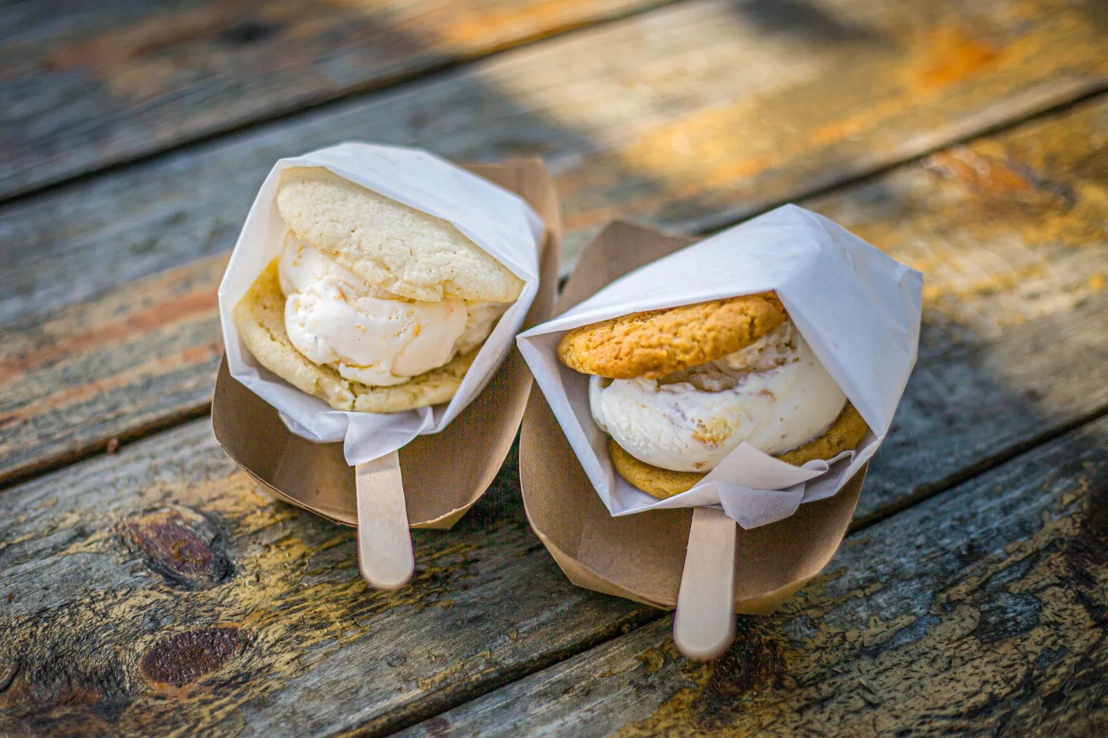 Two ice cream sandwiches in paper wraps on a rustic wooden table outdoors.
