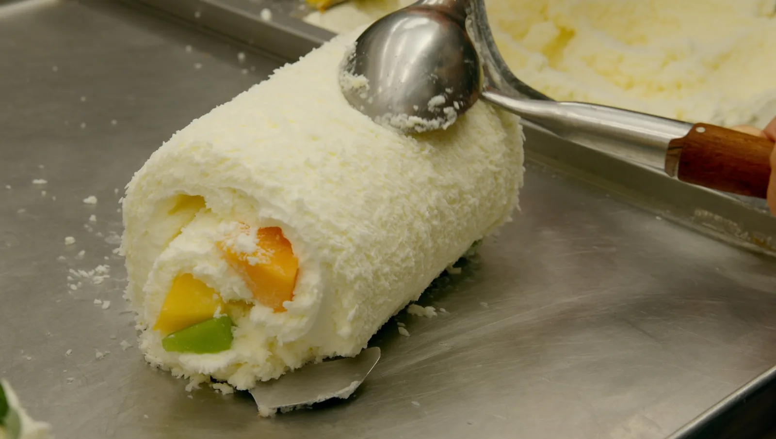 Metal spatulas scraping a creamy coconut base into tight rolls on a frozen steel cold plate.