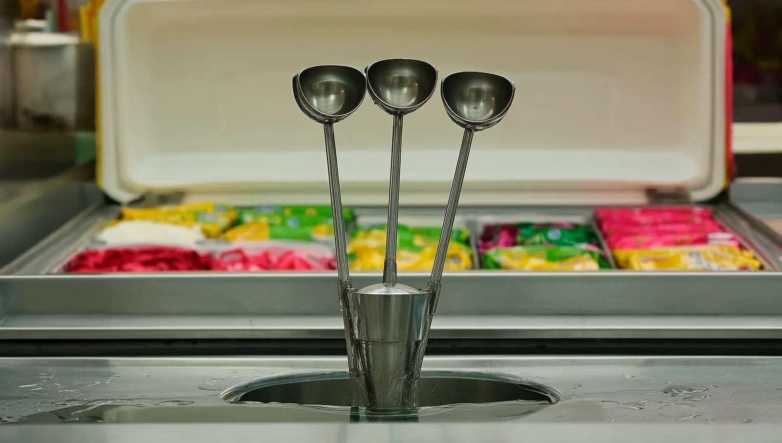 Three metal ice cream scoops standing upright in a stainless steel dipper well of water on a shop counter, with a freezer case behind.
