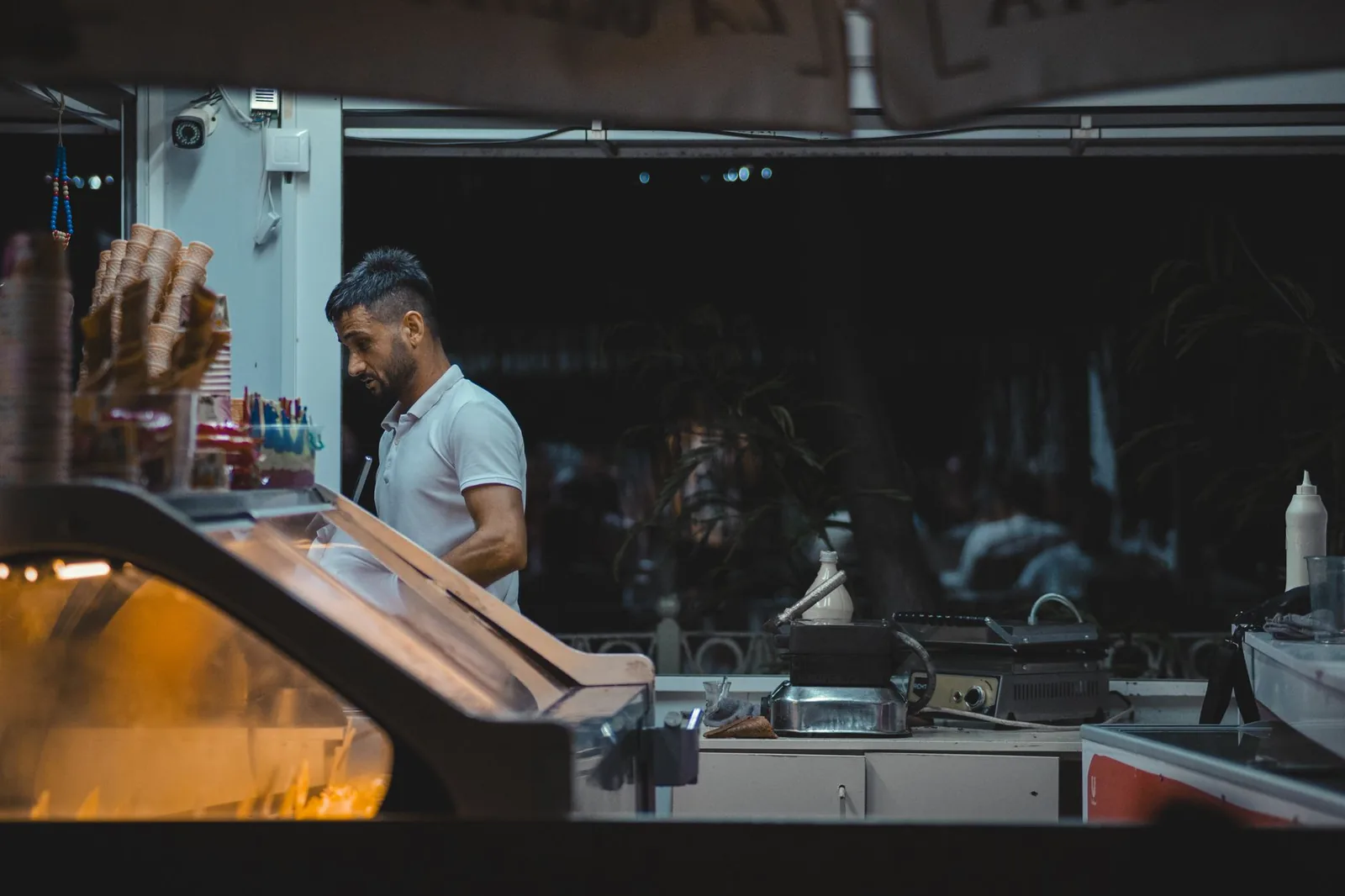 A worker behind the counter of an ice cream shop at night, surrounded by cones and equipment.