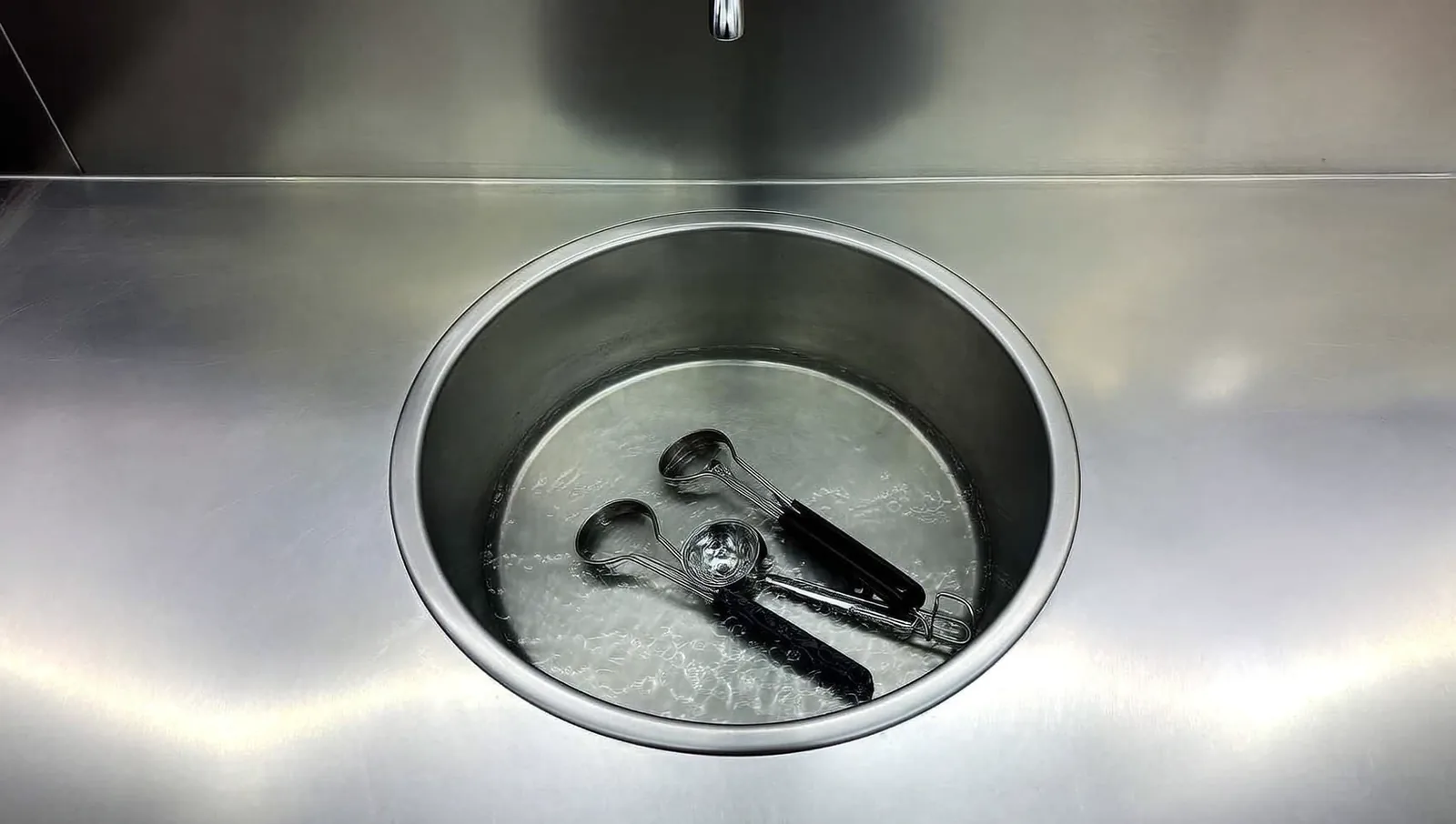 A round stainless steel dipper well of running water set into a bare steel scoop shop counter, with metal ice cream scoops resting inside it