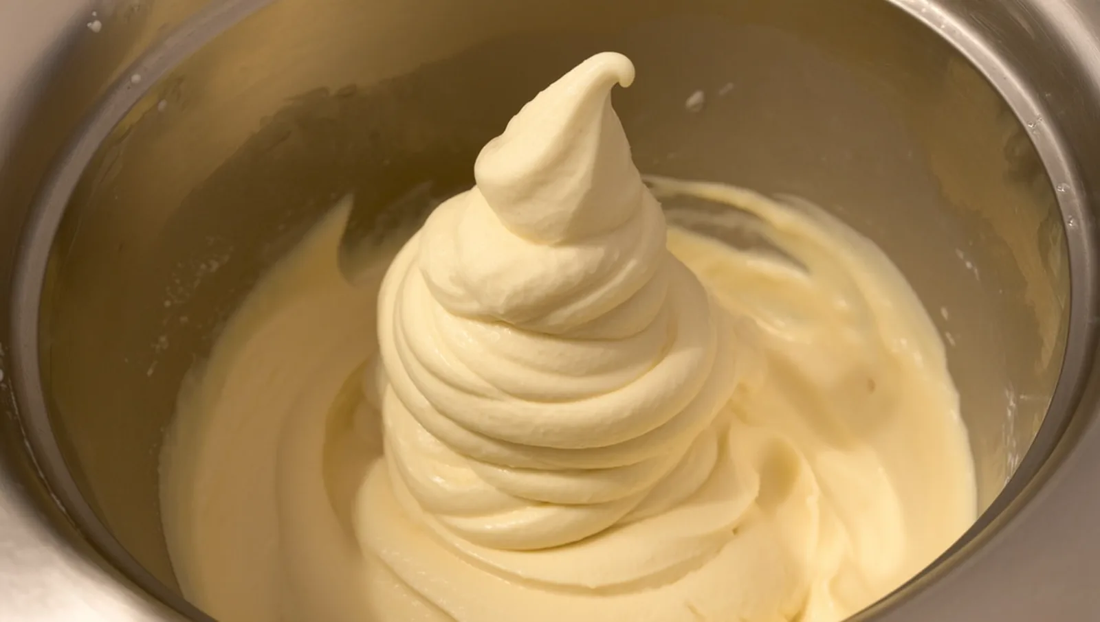 Pale coconut ice cream base churning into a soft-serve swirl inside the metal bowl of a home ice cream maker.