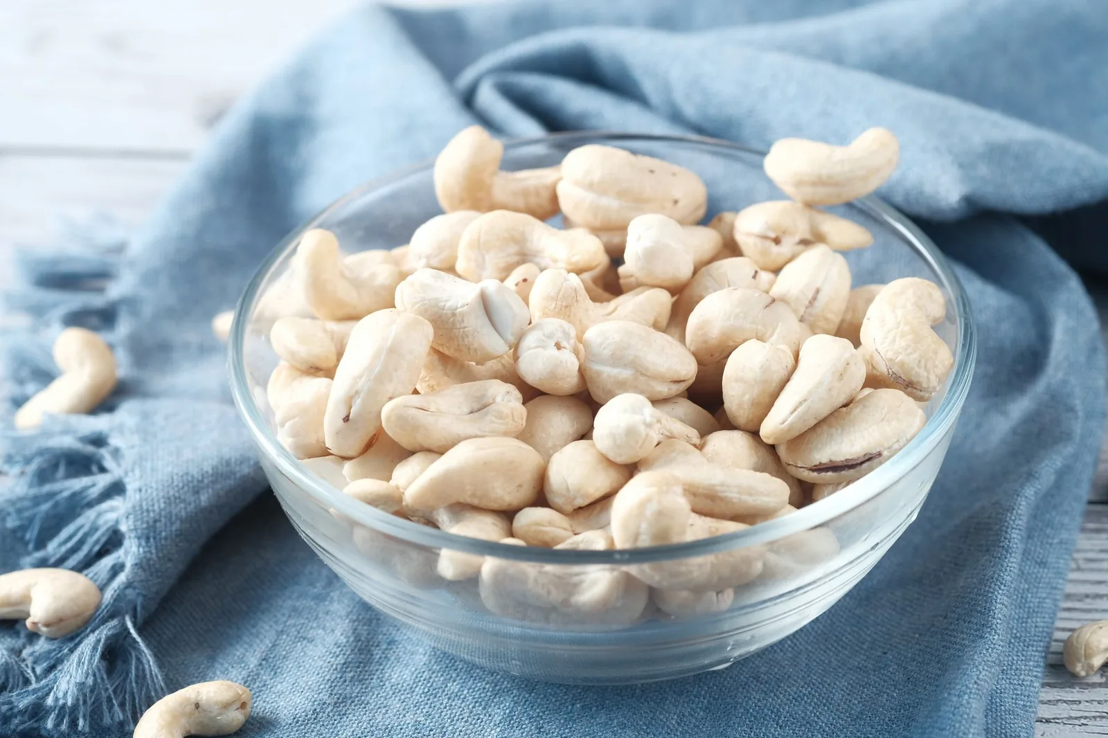 Raw cashews in a clear glass bowl on a blue cloth.