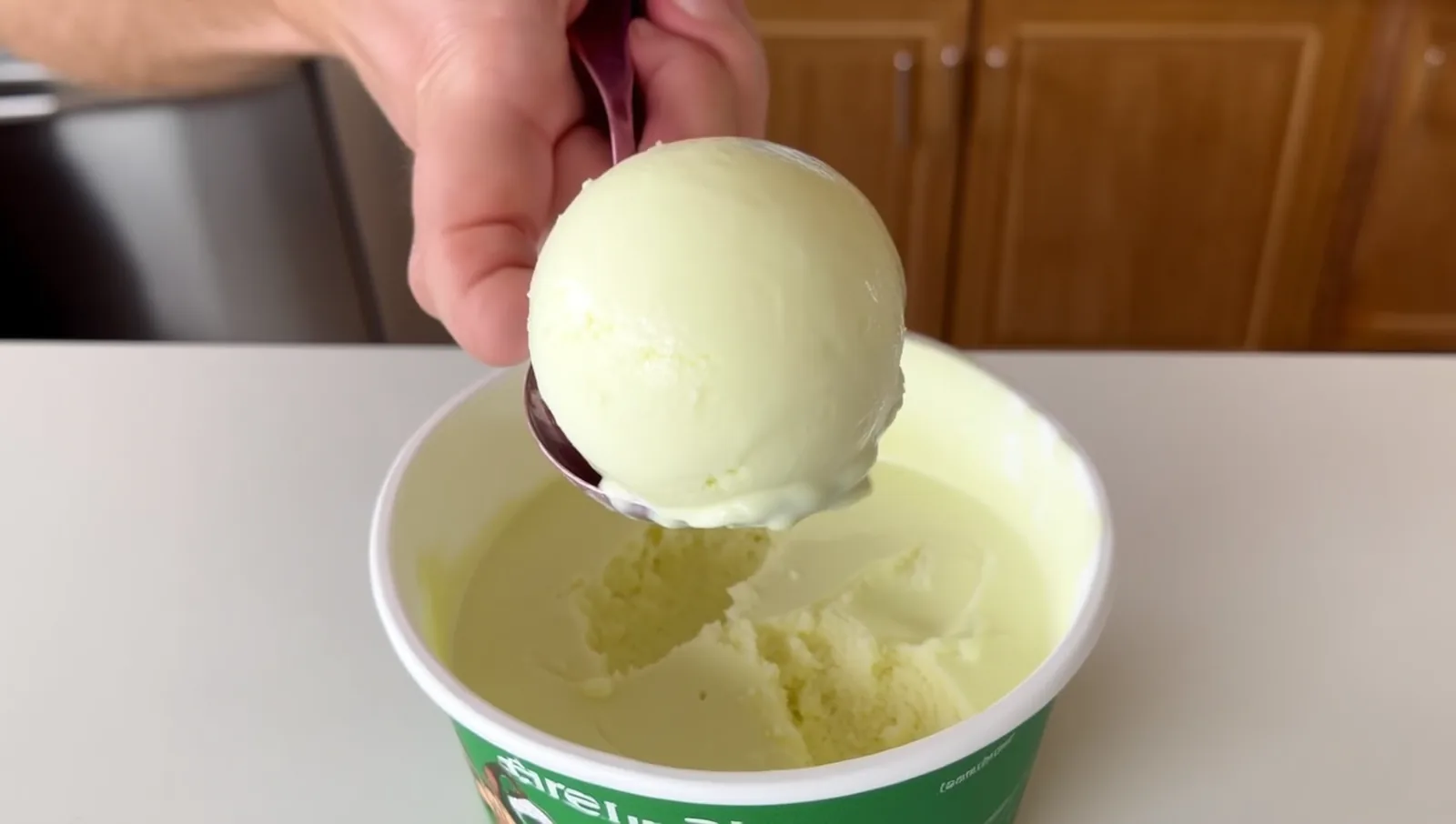 A hand lifting a soft scoop of pale dairy-free ice cream from a freshly churned tub in a home kitchen.